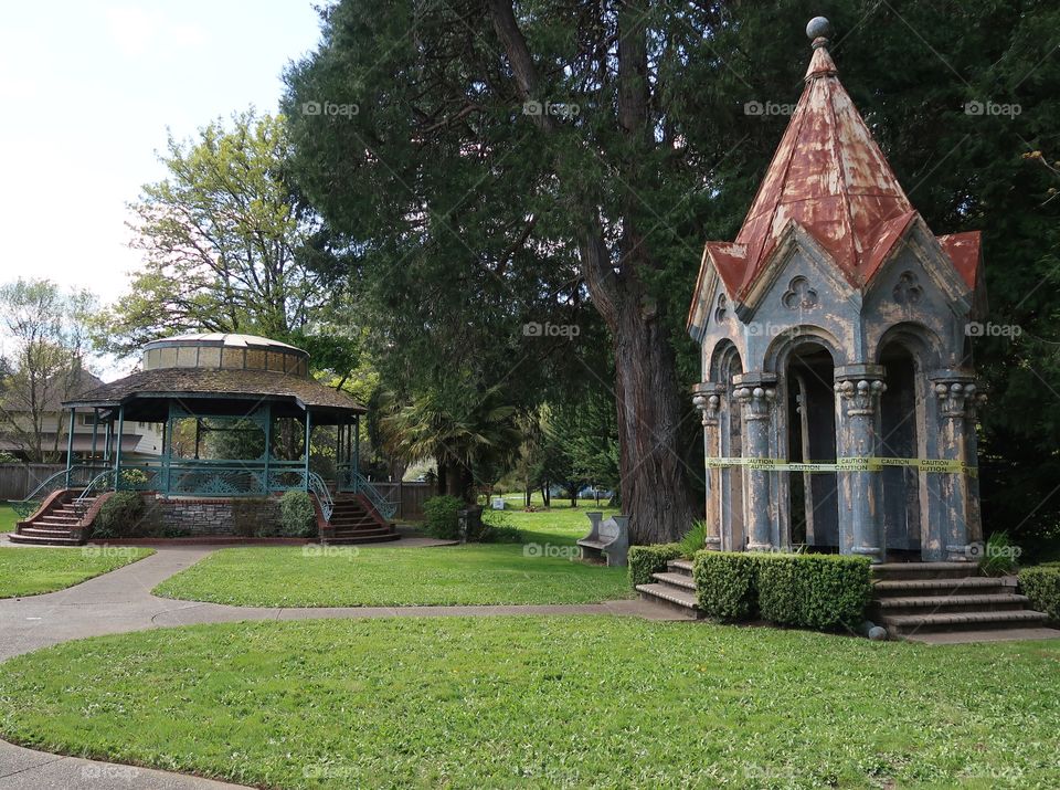 A large decorative stone gazebo in a nice park that has developed a lean and is in need of repair. 