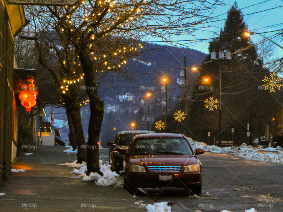 Shot of lights at dusk along a small town street. Red Chinese lanterns at a restaurant, golden hued Christmas & streetlights reflect off sidewalks, cars, road & the snow. Blue hued mountains & a darkening sky as the sun sets behind the mountains.