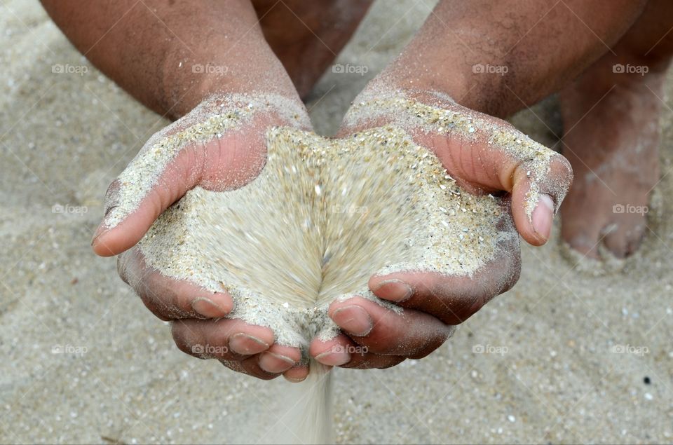 A man squatting on the beach has sand pouring through his hands.