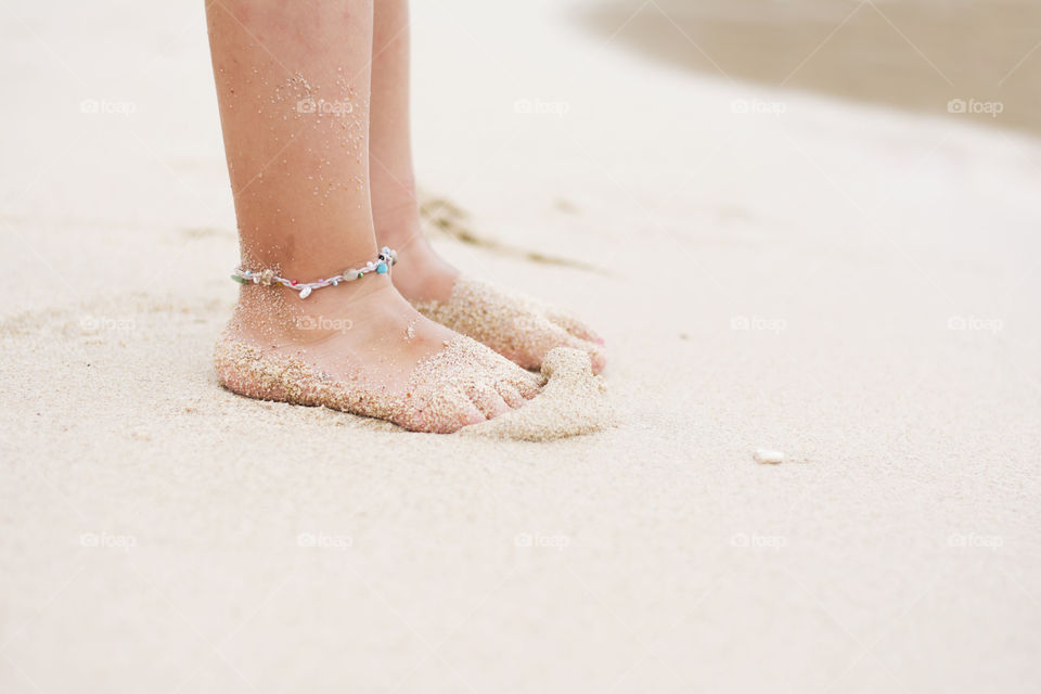child feet with ankle bracelet. little girl standing barefoot on sandy beach near water and wearing ankle bracelet