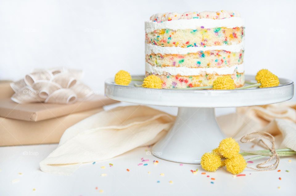 Light and bright image of a colorful confetti layered cake on a cake stand with flowers and gifts