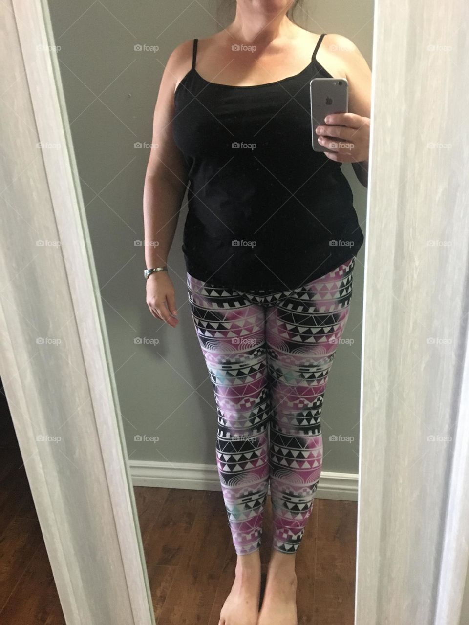 A plus sized, middle aged white lady, models her new black tank top, with her old pink, black, white and blue patterned leggings, showing her bare ankles, arms and shoulders, with only a wrist watch and her phone as assessories