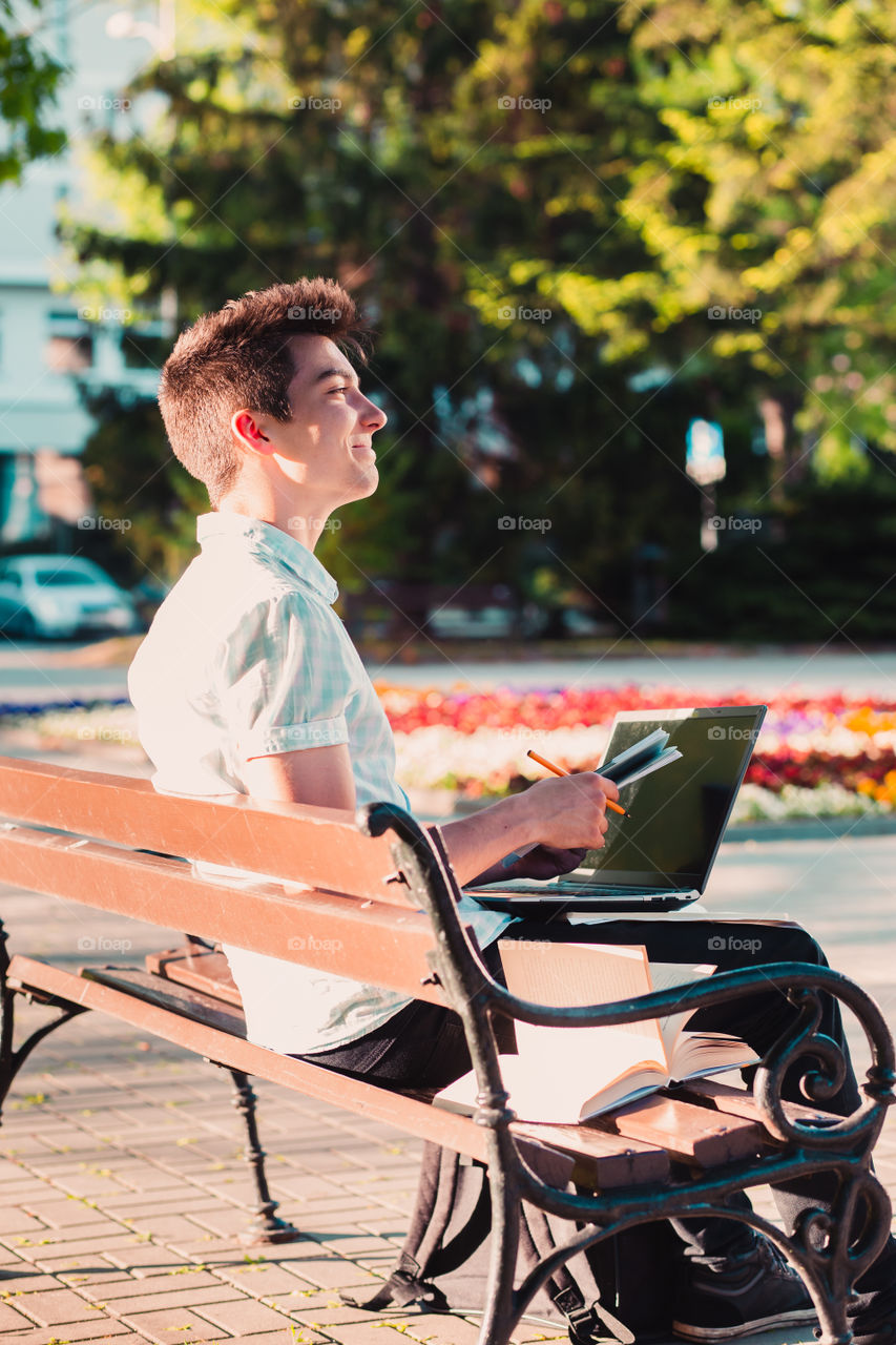 Student working on a laptop using books and notes sitting on a bench in a park. Young boy wearing a blue shirt and dark jeans