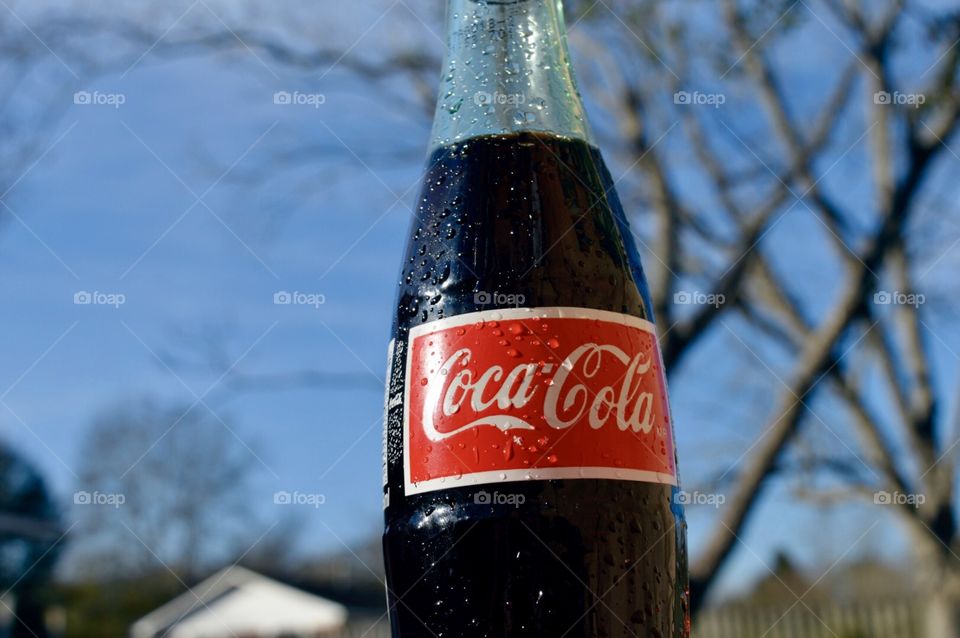 A bottle of Coca-Cola outside with a tree in a rooftop in the background in a subdivision