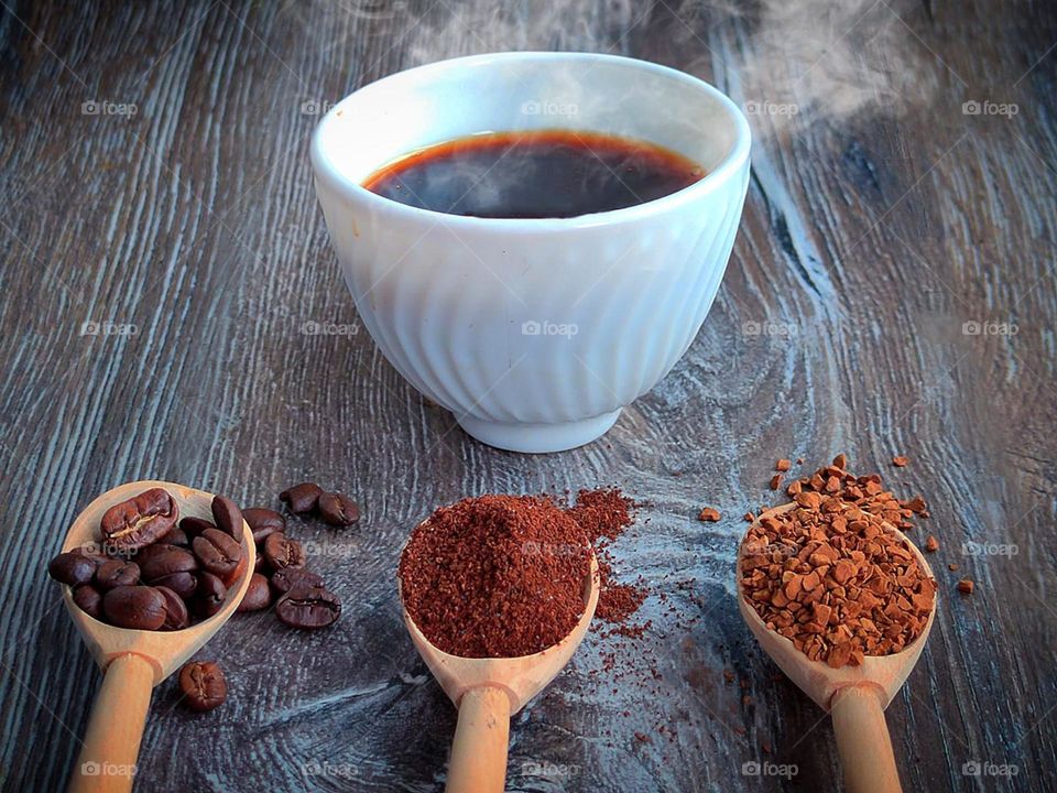 A white cup filled with hot black coffee that steams. In front of the cup are three wooden spoons with different types of coffee: bean coffee, ground coffee and granulated coffee. Wooden background