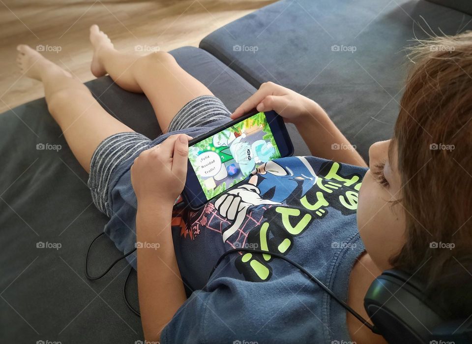 Perspective shot of my 7 year old son on the couch playing a video game on his smartphone with his headphones.