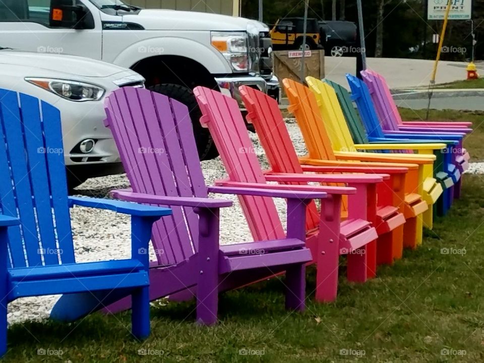 Adirondack chairs for sale along road side, lined up in their rainbow colors. They are all in a row on the grass.