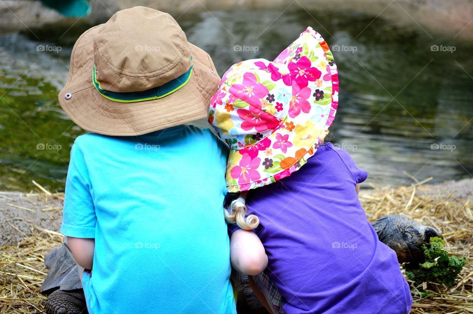 Young siblings wearing sun hats, sitting and snuggling together outdoors with backs to the camera