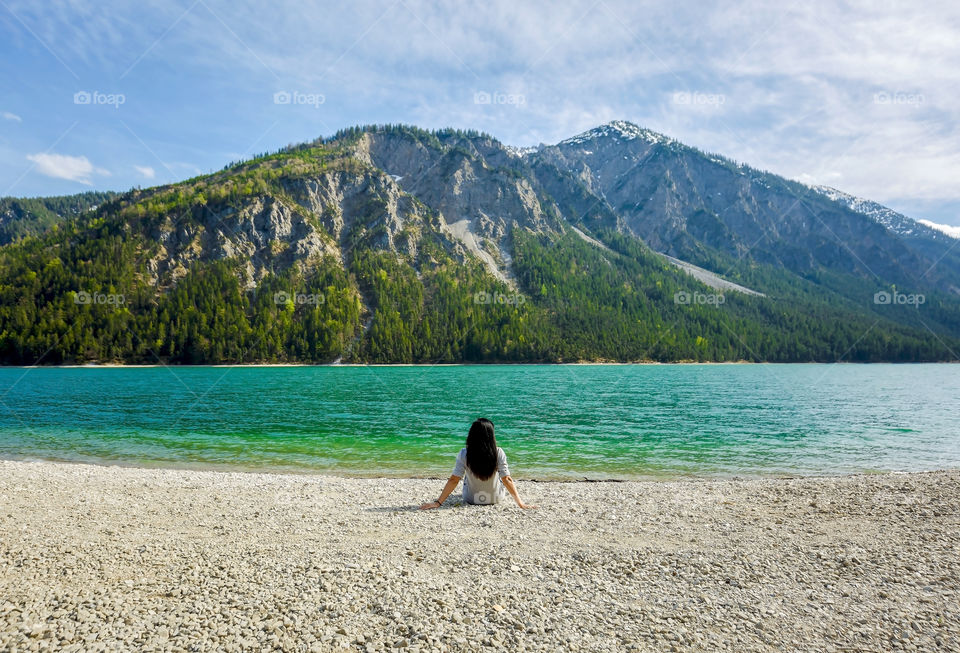 People from behind, woman sitting on lakeside beach with beautiful green water in lake and background of mountains in Austria
