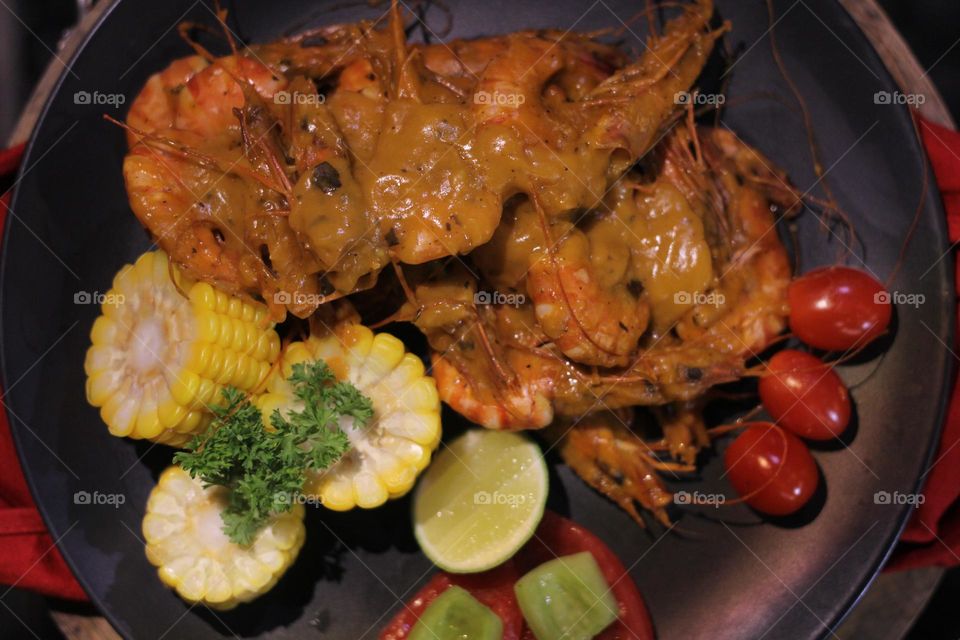 Asian food balado fried prawns with corn chunks, lemon slices and tomatoes served on black plate with red cloth bottom and wooden coaster. Asian style street seafood.