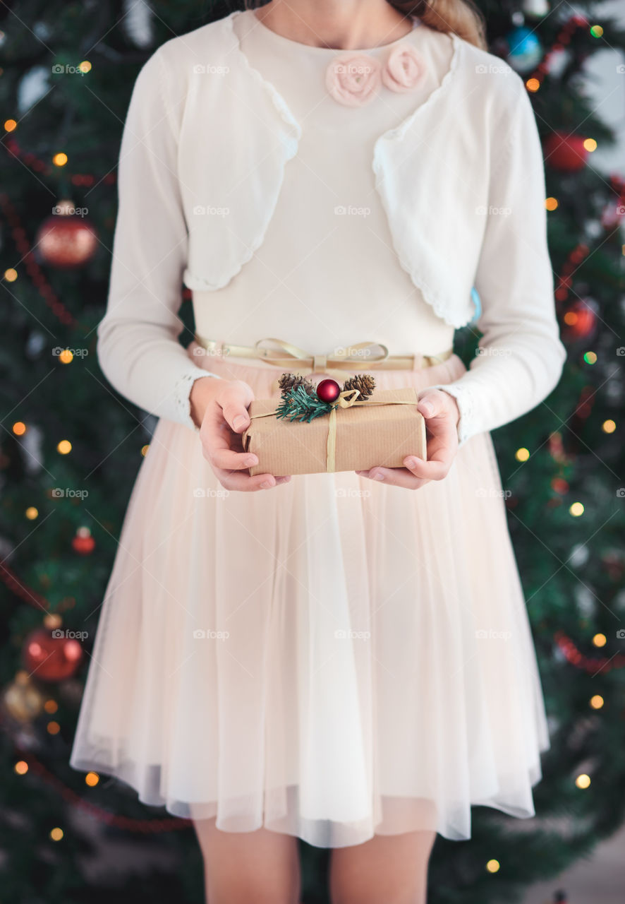 Girl wearing pink dress holding Christmas gift and standing in front of Christmas tree