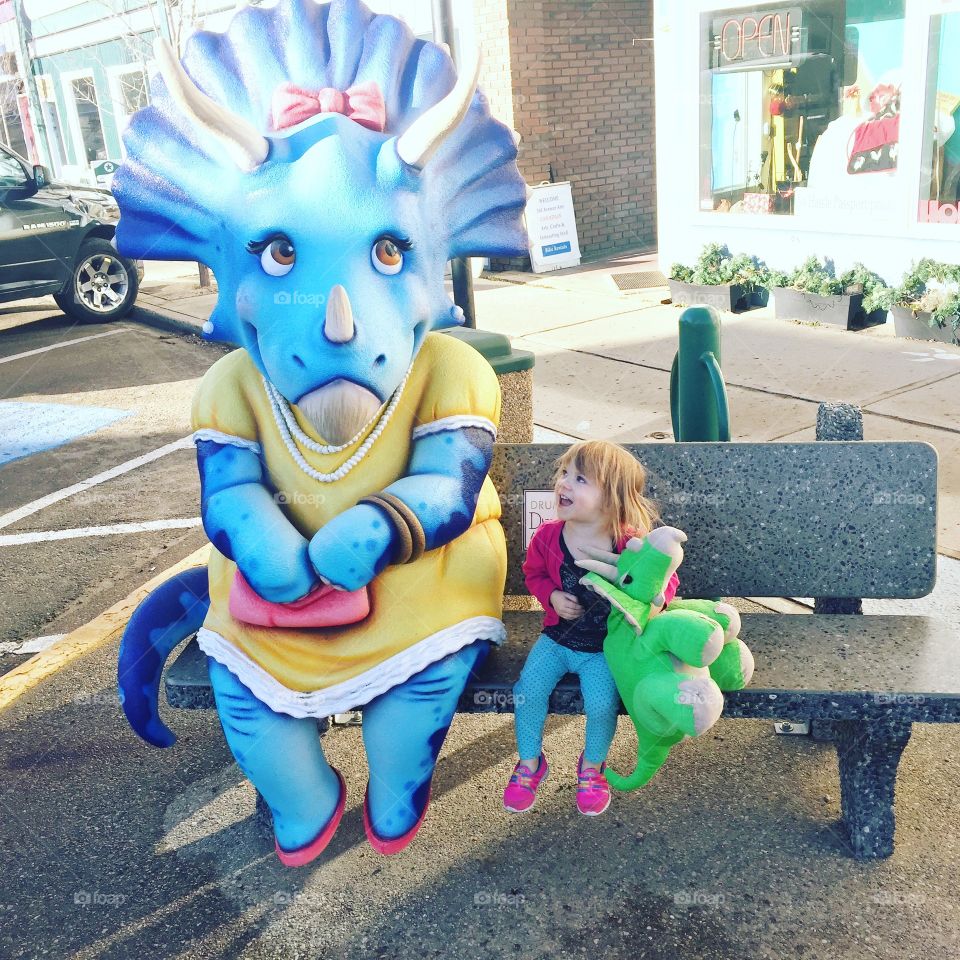 Little 3 year old sitting on a bench with her stuffed triceratops looking fondly at a statue triceratops in Drumheller, Alberta, Canada