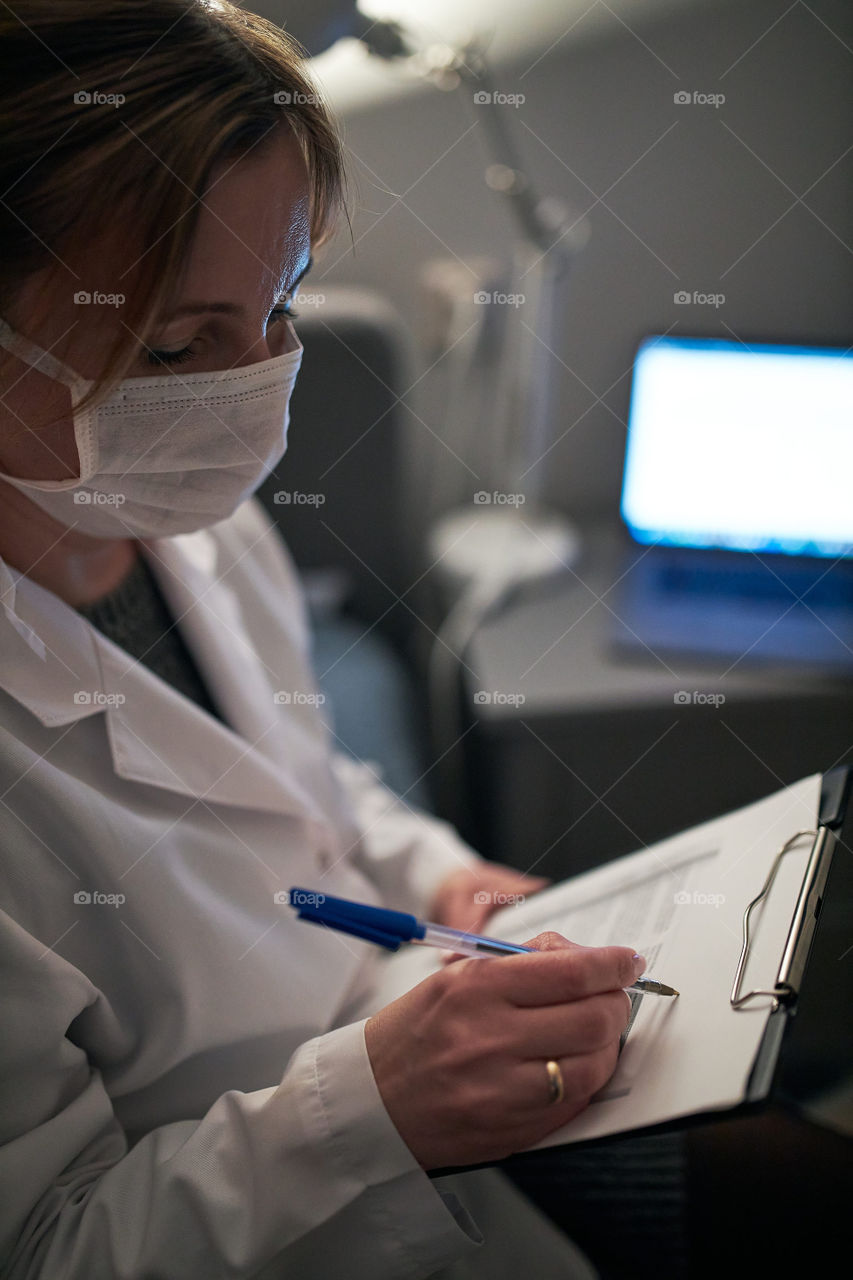 Doctor filling out a document. Hospital staff working at night duty. Woman wearing uniform, cap and face mask to prevent virus infection