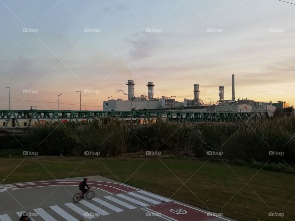 Sun setting behind a factory, a bridge, the train and a cyclist