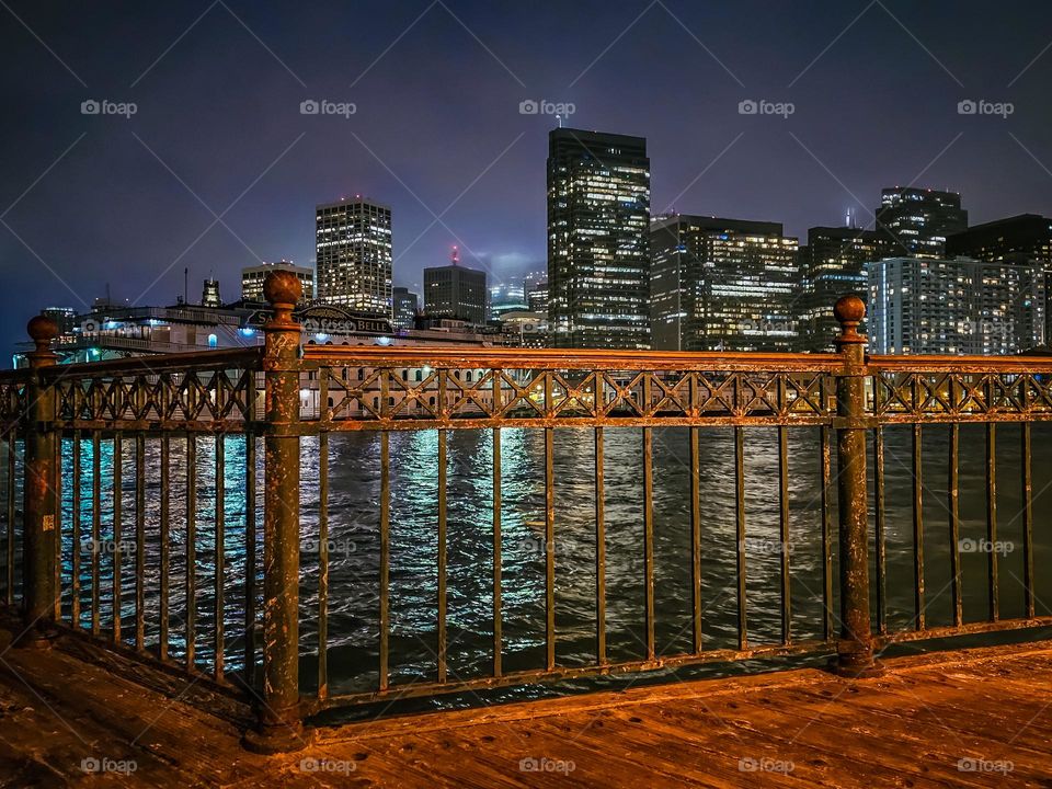 Night view of San Francisco looking through the railing at pier 7 on the waterfront with a slight bit of fog and the lights reflecting in the water 