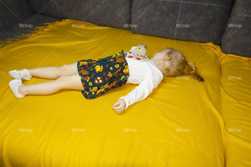 A small, red-haired girl is laughing at home on a gray sofa with purple pillows and a yellow background