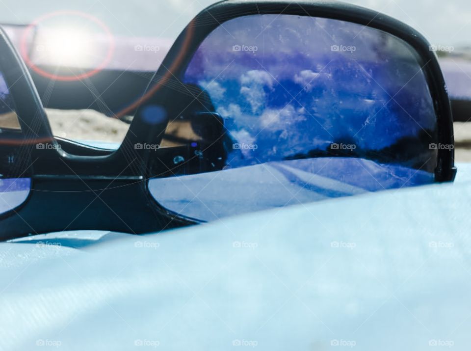Bright blue skies and gorgeous clouds reflected in a pair of sunglasses on the beach.
