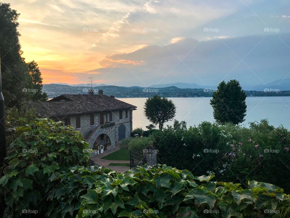 A house by the lake, with a garden, many trees around. Built of stone and the roof was tiled , Iron gate.The house has a beautiful view of the lake, It was surrounded by many mountains, the sky was cloudy and very beautiful in orange.