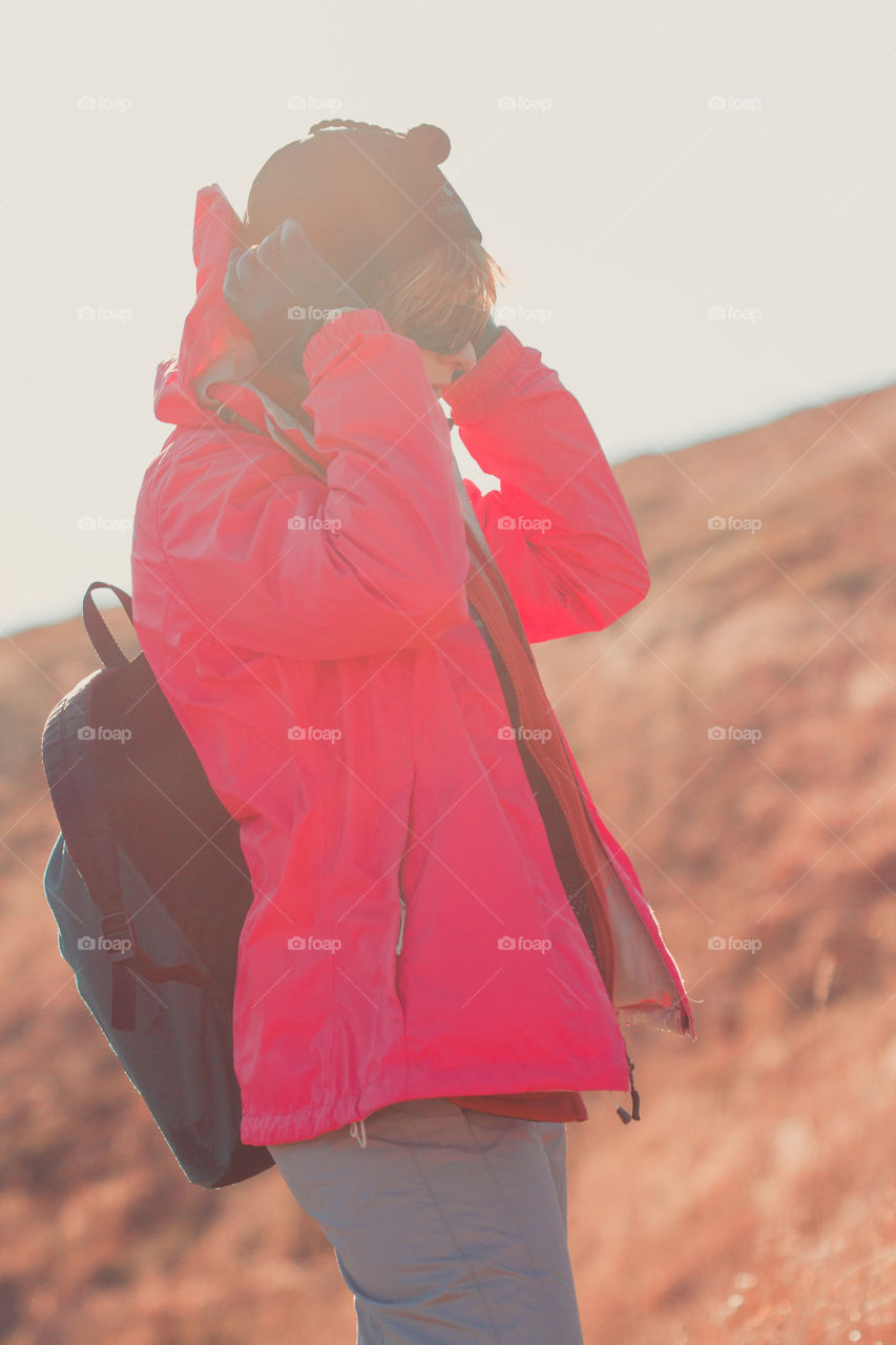 Woman putting a hat on during a trip in the mountains on sunny windy autumn day. Direct sunlight into a lens. Woman wearing sports clothes and carrying backpack