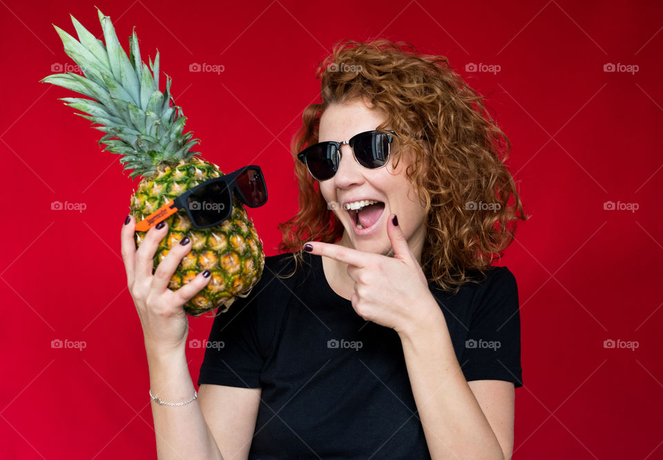 beautiful red-haired girl, in a black T-shirt with a pineapple in her hands, on a red background, has fun and frolics.  Summer mood. Sexy happy girl 