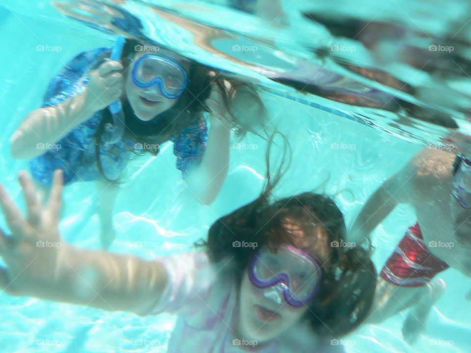 Three persons, dad and two children swimming under the water happy and enjoying having fun in the pool saying hi under the water with goggles.