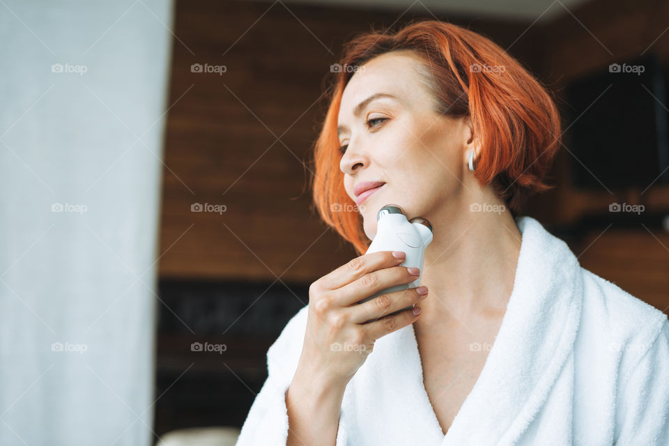 Adult woman with facial microcurrent massager in hands in white bathrobe in bath room spa