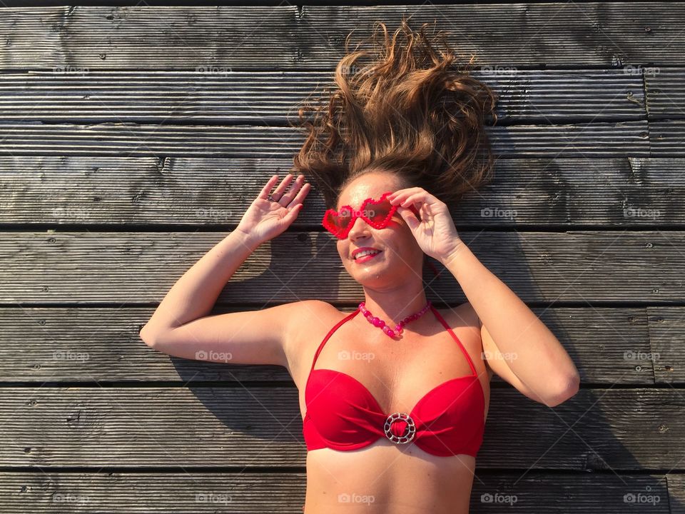 Young woman wearing red bikini and heartshaped sunglasses lying on her back on wooden deck