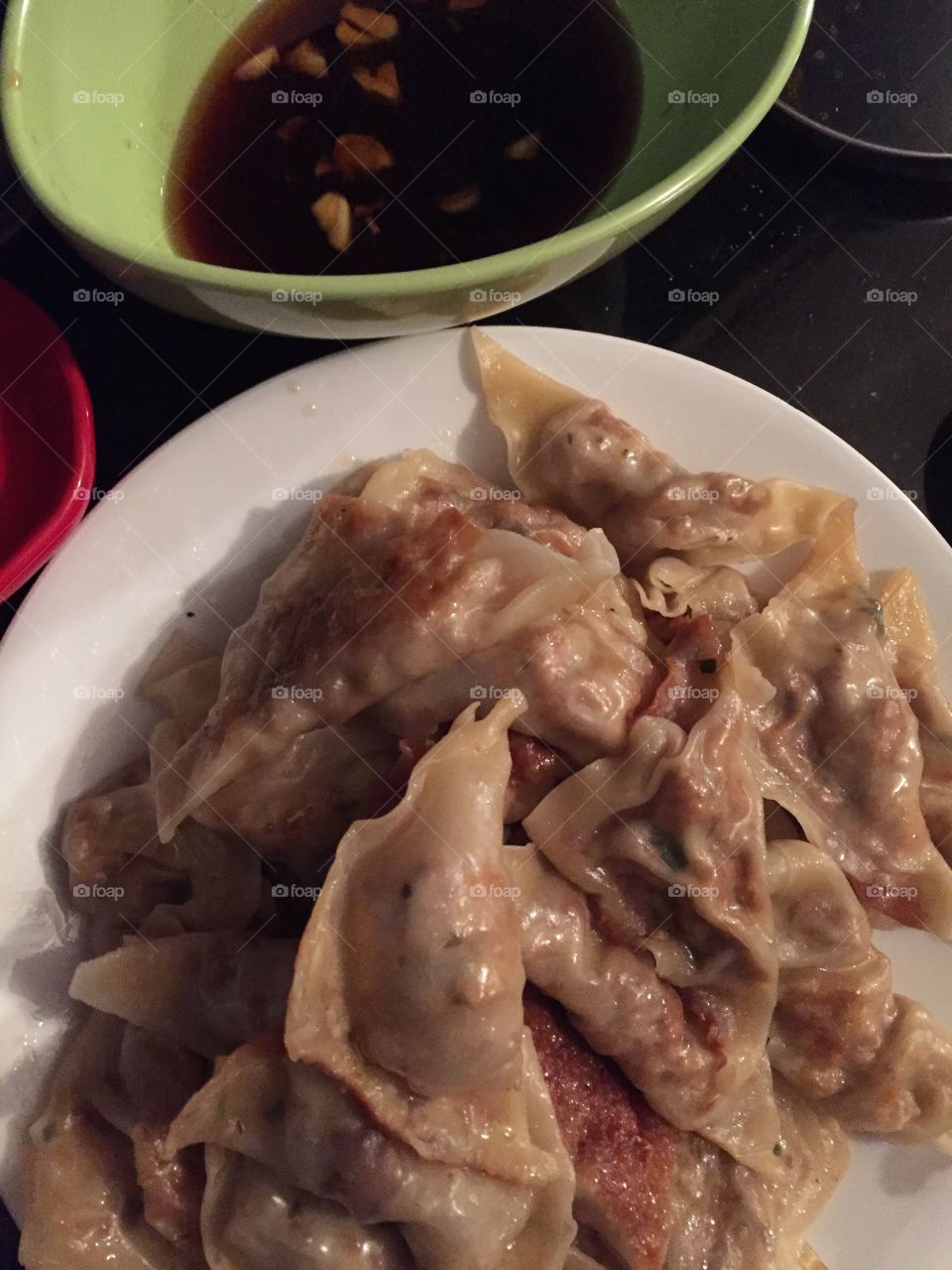 Pile of steamed potstickers on a white plate, next to a green bowl which is filled with garlic infused soy sauce. 