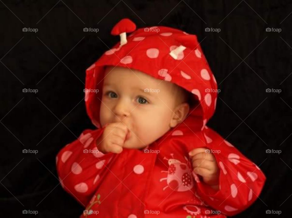little girl in cute red ladybird rain jacket suckling on her thumb 

isolated on black