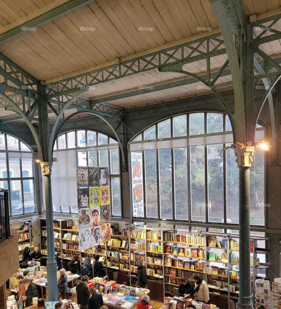 Bookshop full of clients with art ads and stylish architecture with high ceiling and windows.