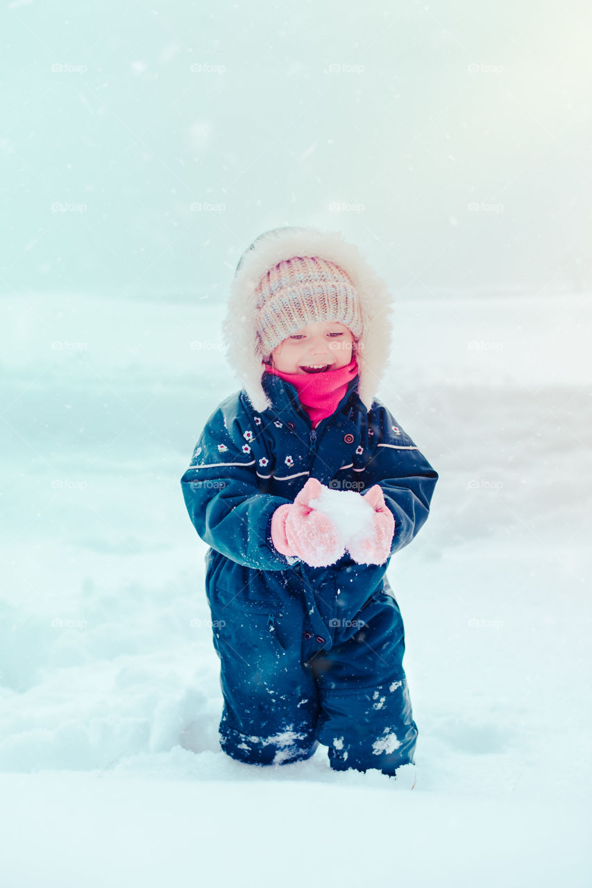 Happy little girl enjoying snow. Child playing outdoors walking through deep snow in wintertime while snow falling. Toddler is wearing dark blue snowsuit
