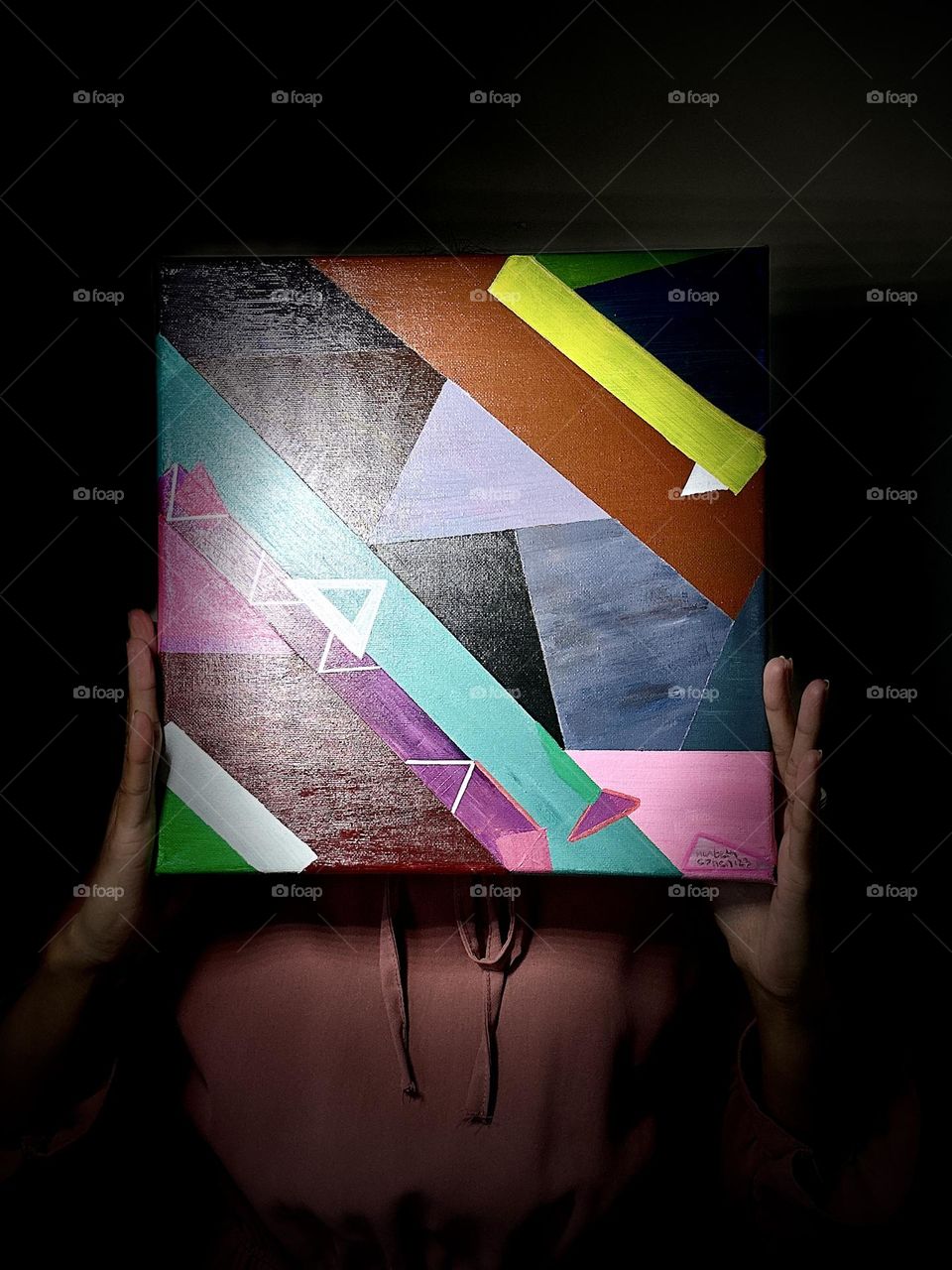 A person’s holds a geometric abstract acrylic painting in front of the face with dark background. 