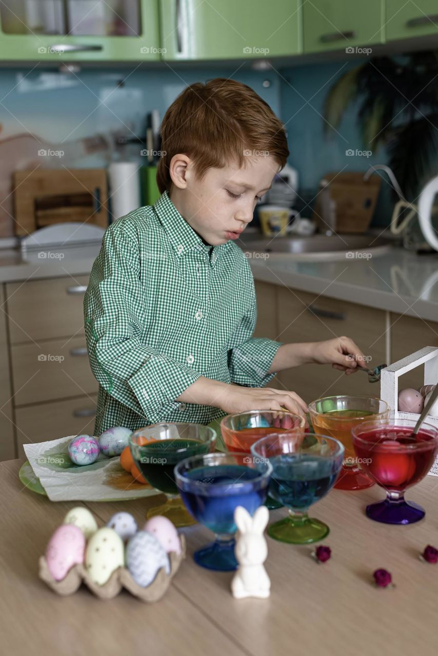 Child red-haired boy paints eggs at home for Easter holiday