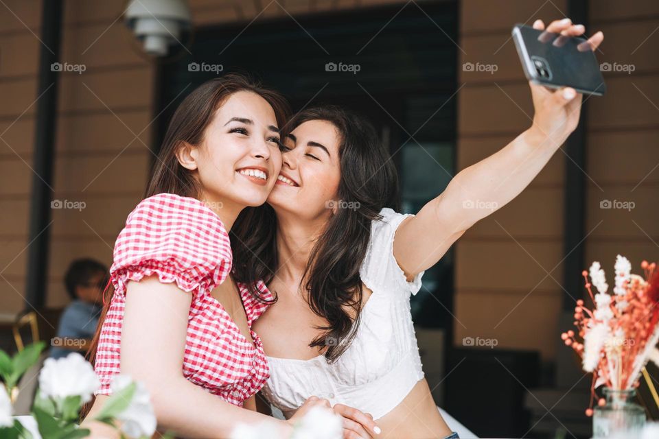 Happy smiling beautiful brunette young women friends in summer clothes taking selfie on mobile phone at the summer city street