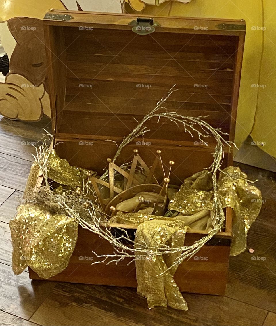 A brown wooden treasure chest with gold ribbon, wreath, and crown inside 