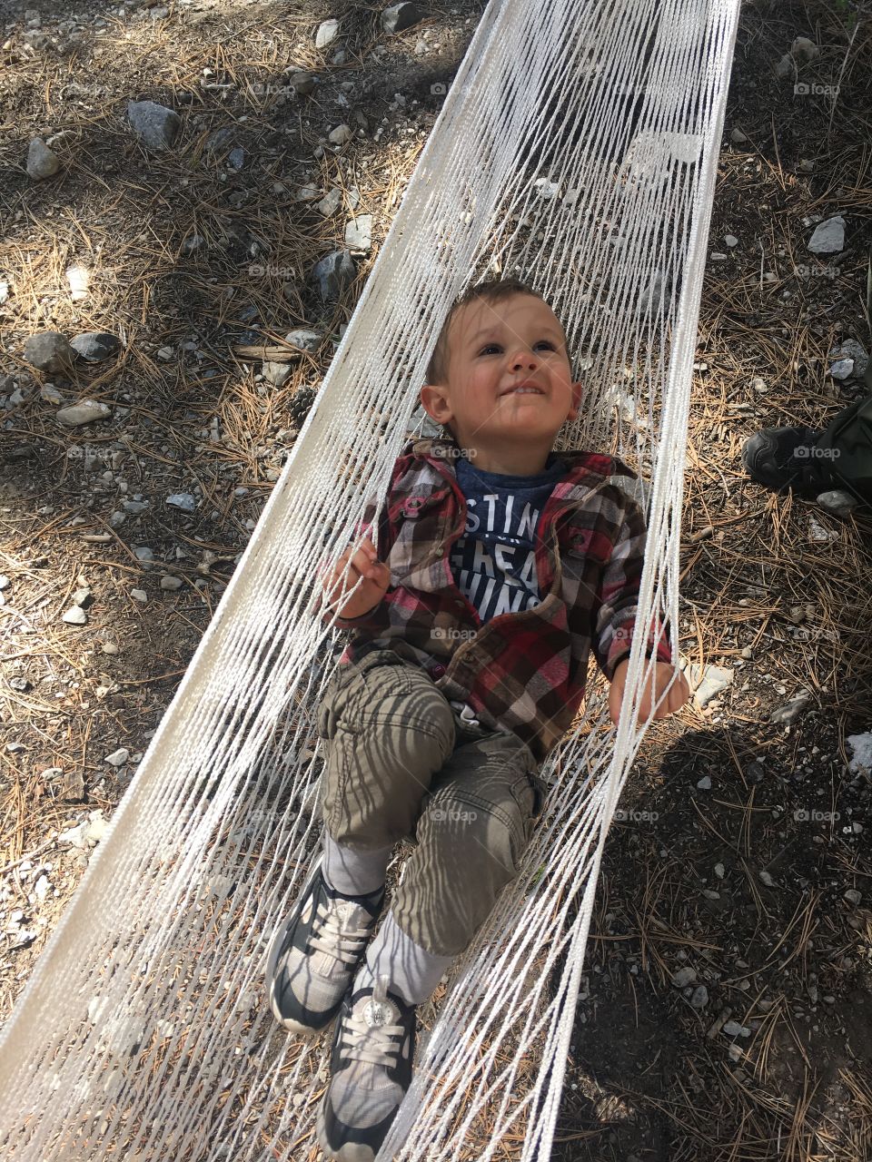An adorable toddler boy wearing a red flannel shirt out camping hanging in a hammock looking up at the sky, smiling.