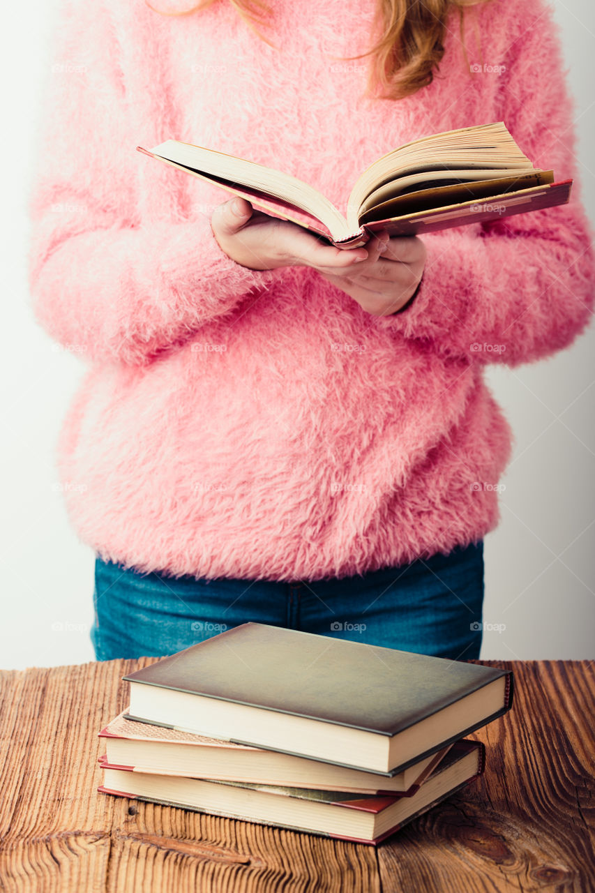Young girl holding a book standing in bookstore. A few books on a wooden table. Teenager girl wearing pink sweater and blue jeans. Vertical photo