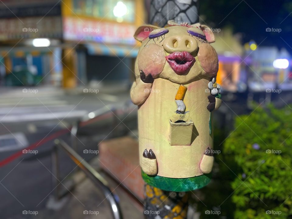 A fancy garbage for cigarettes shaped like a pig. It is nighttime, and there are shops in the background. 