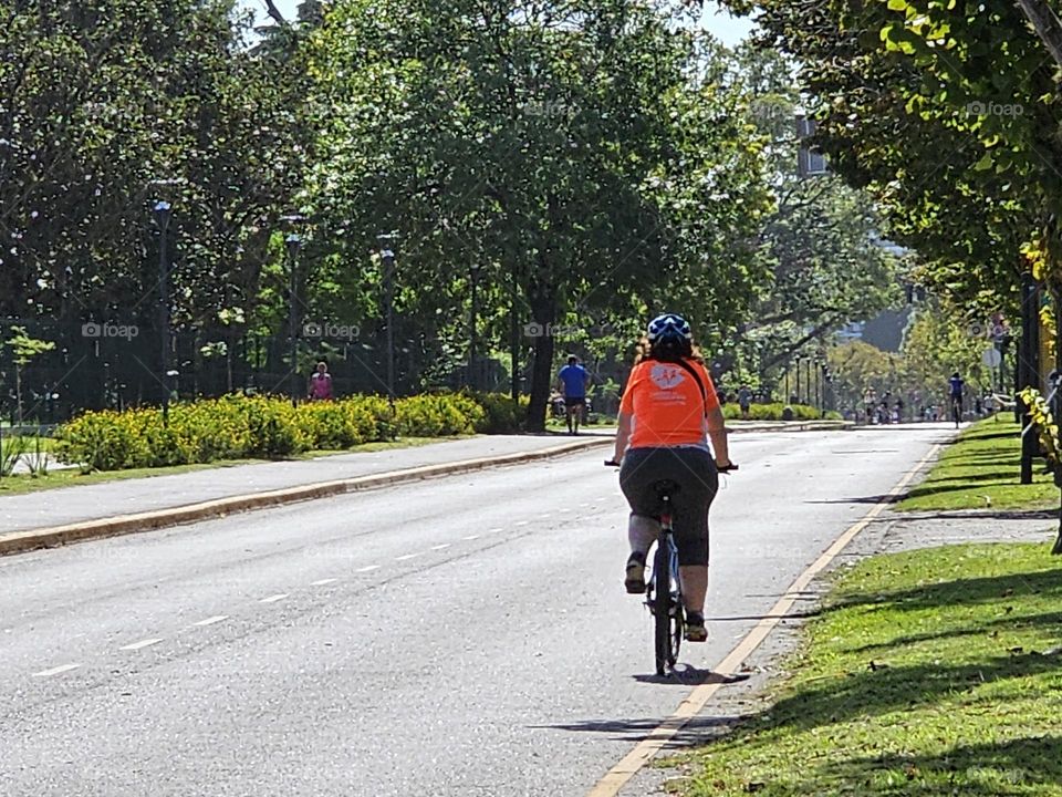 "A lone biker on Sunday". A cyclist with a bright shirt riding on an empty city street, surrounded  by grass on one side, and tall trees on the other. A jogger can be seen at the distance. it's a sunny day. A healthy lifestyle.