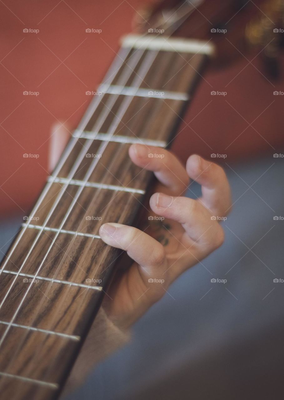 One hand of a little caucasian girl holding a guitar and pinching the strings with her fingers while sitting on the sofa in the room, side view close-up. Music education concept.