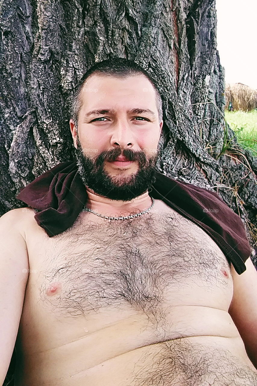 a man with a beard with a bare torso sitting under a tree