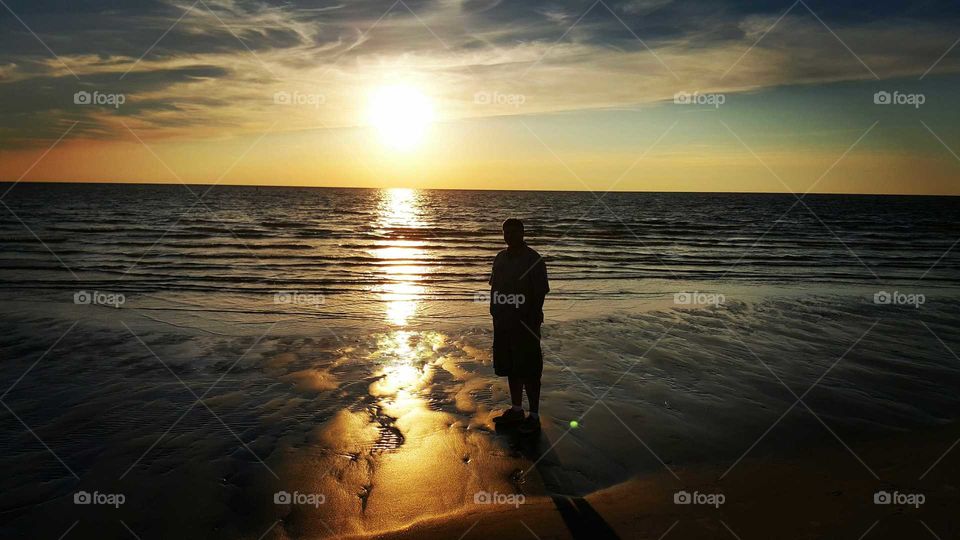 A man in silhouette strolls the beach at sunset reflecting on his day.
