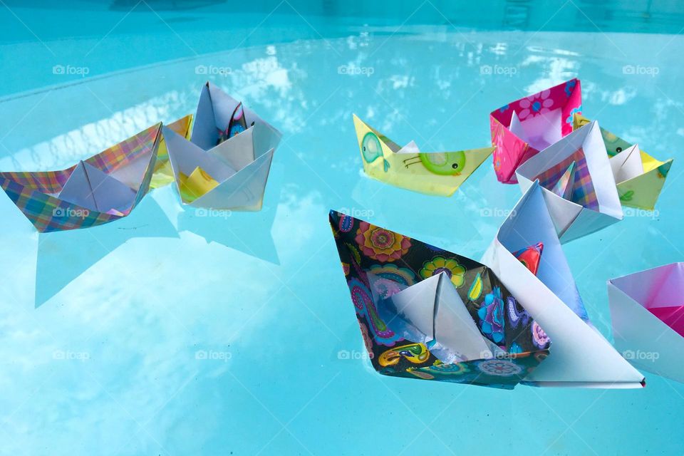 Some colorful paper boats float on the clean blue water of a swimming pool in a sunny day.