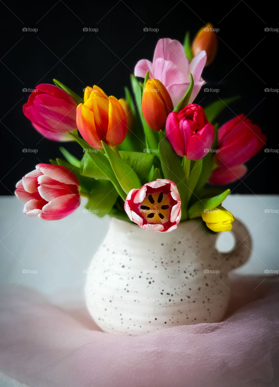 Spring bouquet.  Multi-colored tulips in a white clay jug with a light pink veil on a black background.  Horizontal orientation