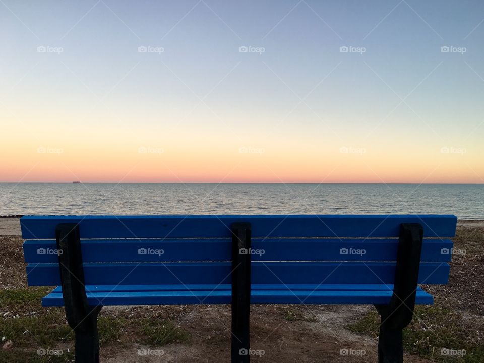 Blue bench foreground on beach at sunset, facing horizon 