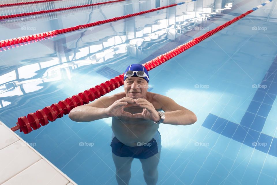 An elderly man, a pensioner in the summer on vacation, in a hotel, stands in a pool with clean and blue water and shows a heart symbol with his hands.