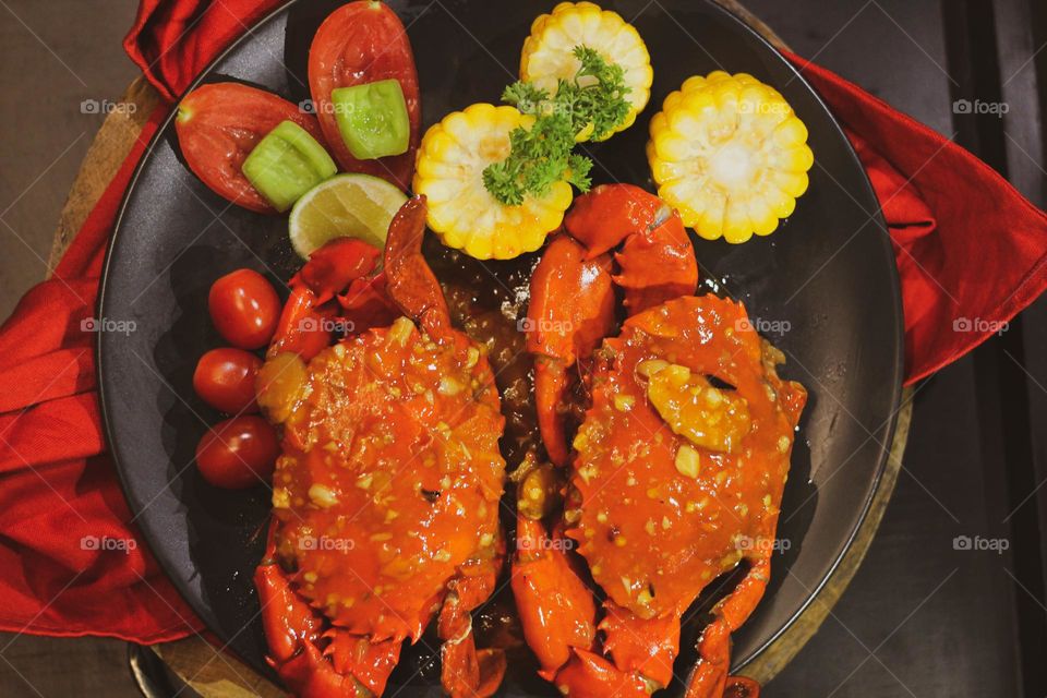 Crab flavored in creamy garlic sauce served with corn, tomatoes, lemon wedges and cherries on a black plate set on a red cloth set in a wooden setting. Asian flavor crab.