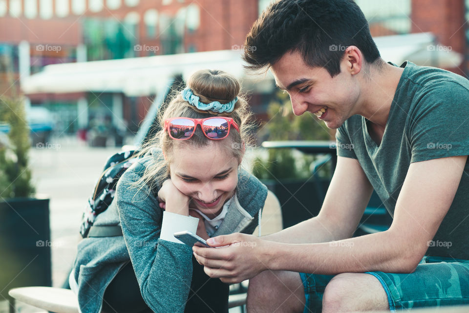 Couple of friends, teenage girl and boy, having fun with smartphones, sitting in center of town, spending time together