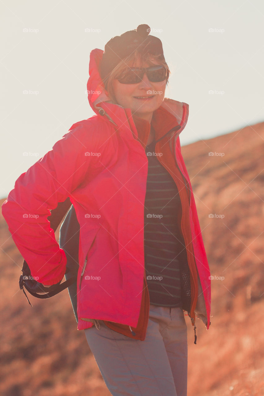 Woman putting a backpack on during a trip in the mountains on sunny windy autumn day. Direct sunlight into a lens. Woman wearing sports clothes smiling and looking at the camera