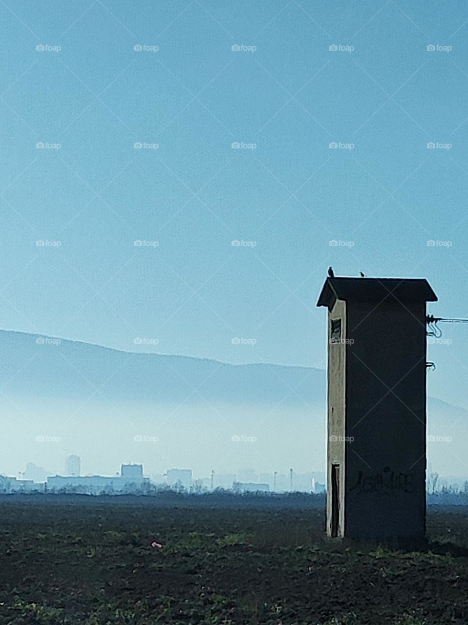 An empty field with foggy hills as a background and a power tower on the front that has two birds sitting on top of it and wires going to the side